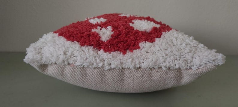K&ouml;nnte beinhalten: Ein dekoratives kissenf&ouml;rmiges Kissen in Pilzform mit einem rot-wei&szlig;en getufteten Design. Die Oberseite des Kissens hat ein rotes Muster mit wei&szlig;en Akzenten, w&auml;hrend die R&auml;nder wei&szlig; sind. Das Kissen steht auf einer neutralfarbenen Stoffbasis.
