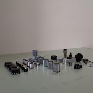 Op de afbeelding: Een verzameling zilveren en zwarte knoppen en andere kleine metalen onderdelen. De onderdelen zijn gerangschikt op een lichtgroen oppervlak.