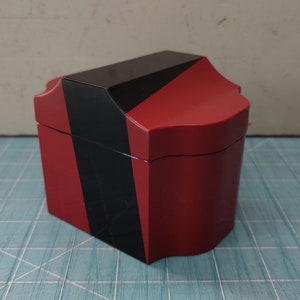 May include: A small, red box with a black geometric design. The box has a hinged lid and a decorative, scalloped edge.