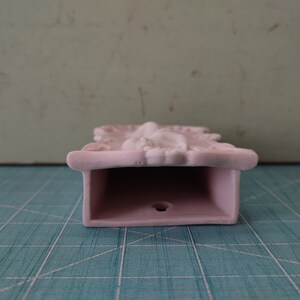 Puede incluir: Un bolsillo de pared de cer&aacute;mica rosa con un dise&ntilde;o ornamentado. El bolsillo tiene una abertura rectangular y un peque&ntilde;o agujero en la parte inferior.