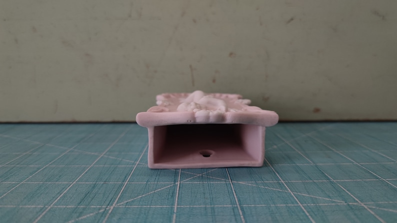 Puede incluir: Un bolsillo de pared de cer&aacute;mica rosa con un dise&ntilde;o ornamentado. El bolsillo tiene una abertura rectangular y un peque&ntilde;o agujero en la parte inferior.