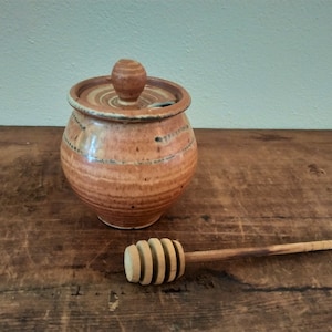 May include: A brown ceramic honey pot with a lid and a wooden honey dipper. The pot has a rustic, hand-crafted look with a glaze that creates a mottled effect.