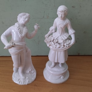以下が含まれることがあります： 白い磁器製の置物2つ。1つはナイフを持った男性、もう1つは花かごを持った女性を表しています。