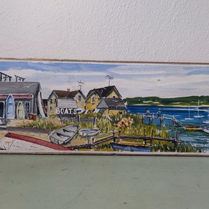 Peut inclure: Peinture rectangulaire d'une scène côtière avec des bateaux et des bâtiments. L'œuvre présente un bâtiment gris avec le mot "BOATS" et une enseigne "Harbor Marine Sales". Plusieurs bateaux sont représentés près du rivage et dans l'eau.