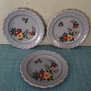 Puede incluir: Tres platos de cerámica blanca con bordes festoneados y ribete azul. Cada plato presenta un diseño floral pintado a mano con flores rojas, amarillas y verdes.