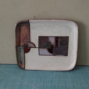 May include: A rectangular ceramic tray with a white background and a geometric design in shades of brown, gray, and pink.