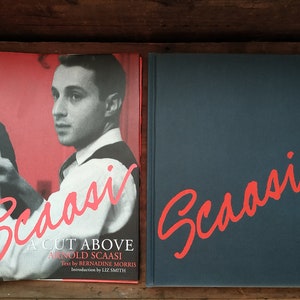 Puede incluir: Un libro titulado "A Cut Above Arnold Scaasi" con un fondo rojo y una foto de Arnold Scaasi en la portada. El libro tiene un título rojo y el nombre del autor está en texto negro. El libro está al lado de un libro azul marino con el título "Scaasi" en texto rojo.