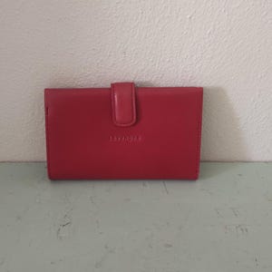 Può includere: Un portafoglio in pelle rosso con chiusura a scatto. Il portafoglio rettangolare ha un piccolo logo in rilievo sul davanti. È appoggiato su una superficie verde chiaro.