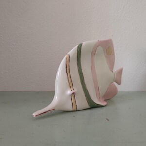 Könnte beinhalten: Eine Keramik-Fischfigur in einem skurrilen Design. Der Fisch ist hauptsächlich weiß mit rosa, grünen und goldenen Akzenten. Er hat einen langen, spitzen Schwanz und einen gebogenen Körper, was ihm ein verspieltes Aussehen verleiht. Die Figur ist ein dekorativer Artikel.