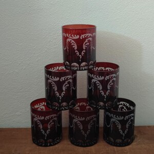 May include: Six red-tinted drinking glasses with a white, ornate, etched design. The glasses are arranged in a pyramid shape on a wooden surface. The glasses are cylindrical and appear to be made of glass.