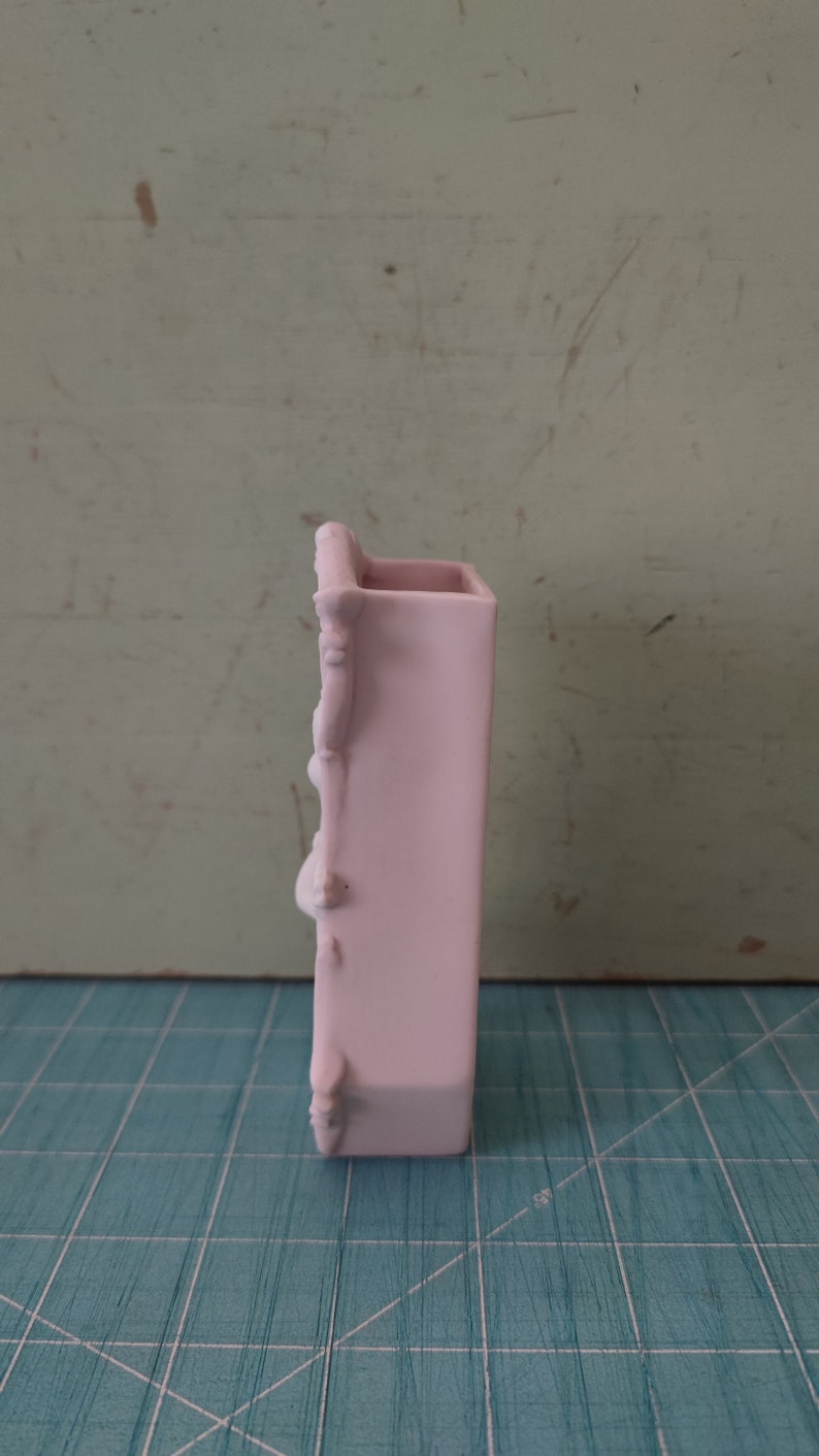 Puede incluir: Un jarr&oacute;n de cer&aacute;mica rosa claro con un dise&ntilde;o decorativo y ornamentado. El jarr&oacute;n es alto y estrecho, con forma rectangular.