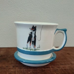 May include: White ceramic planter with a blue band around the top and a blue saucer. The planter features a hand-painted image of a black and tan Basenji dog.