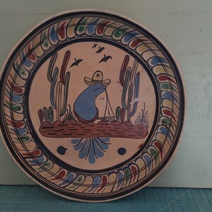 May include: Hand-painted ceramic plate with a desert scene. The plate features a central image of a person in a sombrero, cacti, and birds. The rim has a decorative border with blue, red, and yellow accents. A unique piece of Southwestern decor.