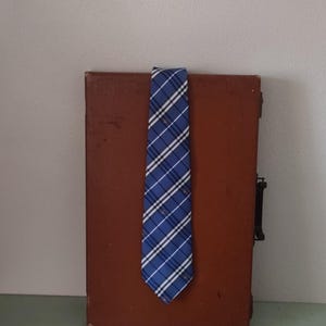 May include: A blue and white plaid tie is draped over a brown, vintage-style briefcase. The tie has a diagonal pattern with thin red stripes. The briefcase is closed and has a black handle and clasp.