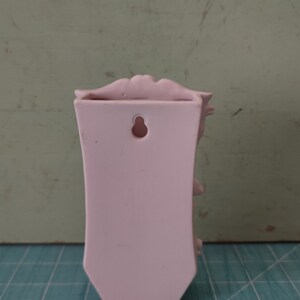 Puede incluir: Un jarr&oacute;n de pared de cer&aacute;mica rosa con un dise&ntilde;o floral decorativo y un agujero para colgar. El jarr&oacute;n tiene una parte superior festoneada y una forma ligeramente curvada.