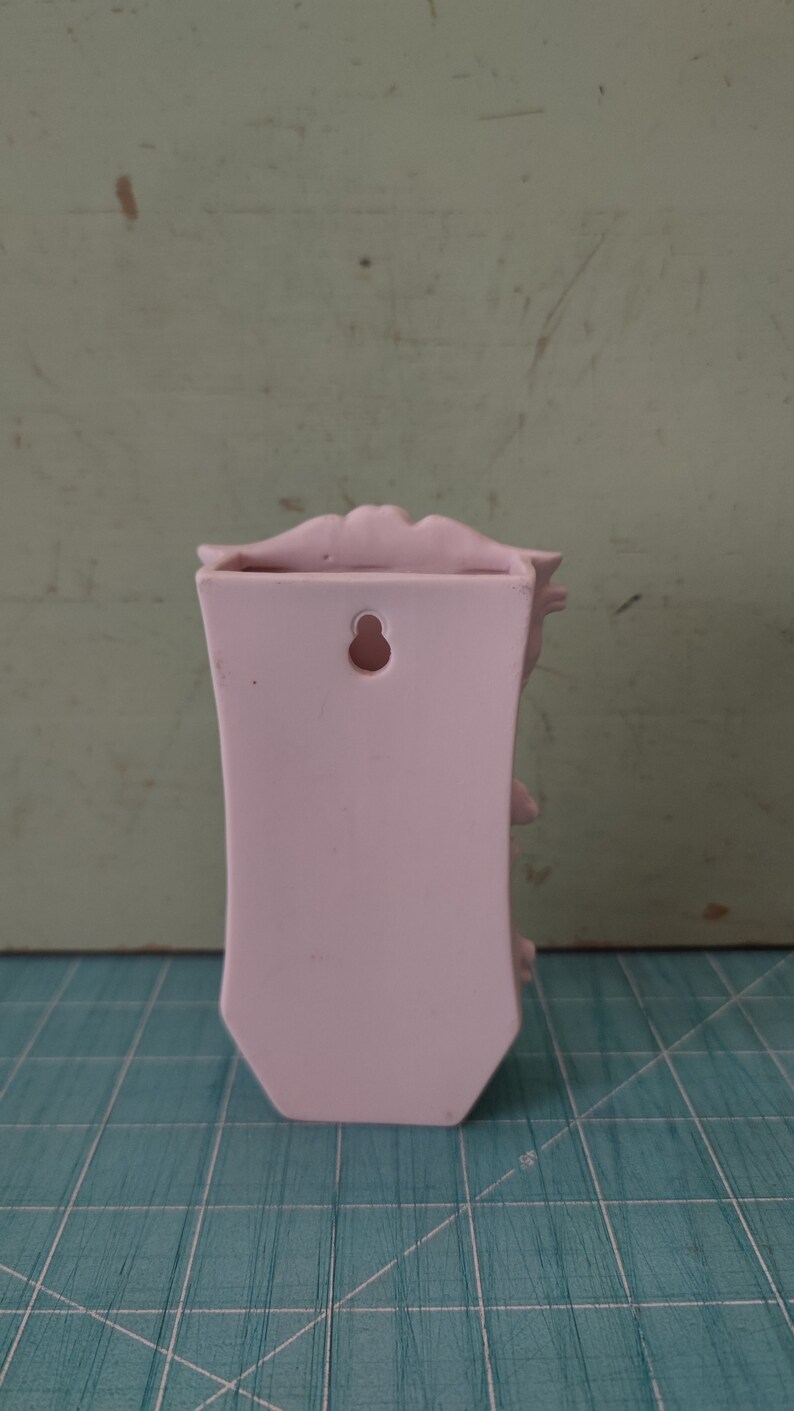 Puede incluir: Un jarr&oacute;n de pared de cer&aacute;mica rosa con un dise&ntilde;o floral decorativo y un agujero para colgar. El jarr&oacute;n tiene una parte superior festoneada y una forma ligeramente curvada.