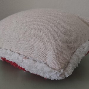 K&ouml;nnte beinhalten: Ein kleines, quadratisches Kissen aus strukturiertem, cremefarbenem Stoff. Das Kissen hat einen wei&szlig;en, gefransten Rand und einen roten Akzent, der am unteren Rand sichtbar ist. Das Kissen liegt auf einer hellgr&uuml;nen Oberfl&auml;che.