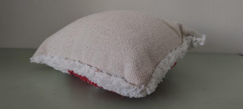 K&ouml;nnte beinhalten: Ein kleines, quadratisches Kissen aus strukturiertem, cremefarbenem Stoff. Das Kissen hat einen wei&szlig;en, gefransten Rand und einen roten Akzent, der am unteren Rand sichtbar ist. Das Kissen liegt auf einer hellgr&uuml;nen Oberfl&auml;che.