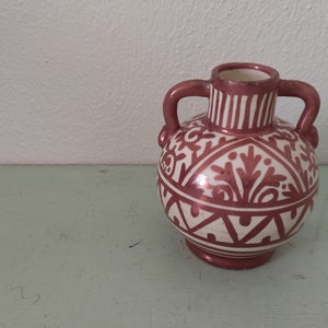 Op de afbeelding: Een kleine, witte keramische vaas met een koperkleurig geometrisch en bloemmotief. De vaas heeft twee handvatten en een rond lichaam.