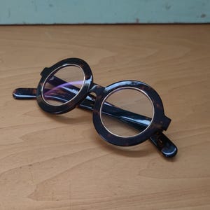 May include: Round-framed eyeglasses with a dark, tortoiseshell pattern. The lenses are clear, and the frames have a thick, bold design. The glasses are resting on a light brown wooden surface.