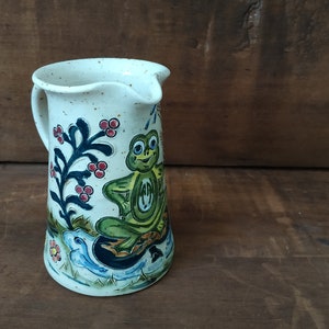 May include: A white ceramic pitcher with a speckled glaze features a hand-painted design of a green frog sitting on a blue and white lily pad. The frog has a red and yellow design on its back and a blue and white flower is behind it.