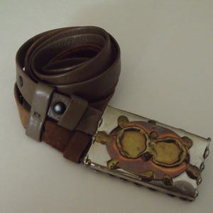 Op de afbeelding: Een bruine leren riem met een decoratieve gesp. De gesp heeft een zilverkleurig frame met koper- en goudkleurige accenten. De riem is opgerold en toont de gesp en de leer- en suède textuur.