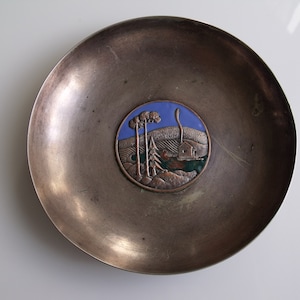 May include: A silver metal dish with a round, raised, copper-colored design in the center. The design depicts a landscape with three trees, a small house, and a blue sky.