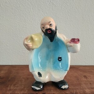May include: A ceramic figurine of a man with a beard and a mustache, wearing a blue and white robe. He is holding a yellow hat in one hand and a red object in the other.