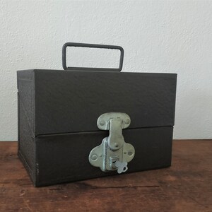 Könnte beinhalten: Eine dunkelgraue Metallbox mit einem Tragegriff und einem silbernen Schloss und Schlüssel. Die Box steht auf einer Holzoberfläche.