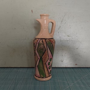 Op de afbeelding: Een kleine keramische kan met een geometrisch ontwerp in tinten groen, roze en bruin. De kan heeft een handvat en een tuit.