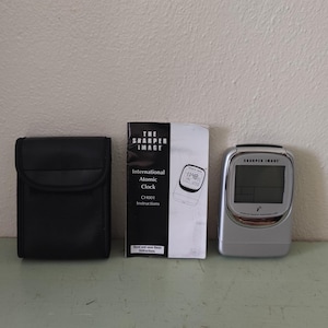 May include: A silver atomic clock with a black carrying case and instruction manual. The clock has a digital display and the text "The Sharper Image" on the front. The manual has the text "International Atomic Clock" and "Sharper Image" on the front.