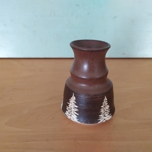 May include: A small brown ceramic vase with a white glaze design of stylized pine trees. The vase has a flared base and a narrow neck.