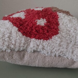 K&ouml;nnte beinhalten: Ein dekoratives Kissen mit einem strukturierten, getufteten Design. Das Kissen hat ein zentrales rotes Blumenmuster, das von wei&szlig;en und beigen Tufting umgeben ist. Die Basis des Kissens ist ein neutraler beiger Stoff.