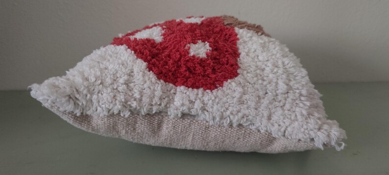 K&ouml;nnte beinhalten: Ein dekoratives Kissen mit einem strukturierten, getufteten Design. Das Kissen hat ein zentrales rotes Blumenmuster, das von wei&szlig;en und beigen Tufting umgeben ist. Die Basis des Kissens ist ein neutraler beiger Stoff.