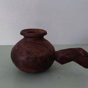 Puede incluir: Una olla de madera marrón oscuro con un cuerpo redondeado y un asa con forma de animal estilizado. La olla tiene un diseño tallado en la superficie.