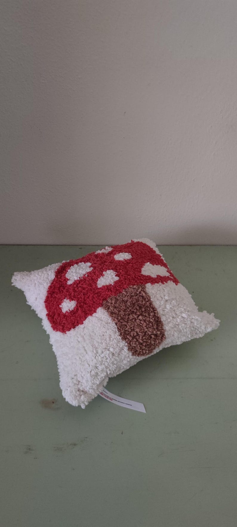 K&ouml;nnte beinhalten: Ein dekoratives Kissen mit einem Pilzdesign. Der Pilzhut ist rot mit wei&szlig;en Punkten und der Stiel ist braun. Das Kissen hat einen wei&szlig;en, strukturierten Hintergrund.