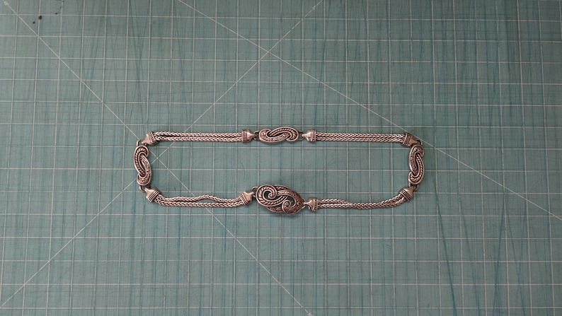 Peut inclure: Un collier en cha&icirc;ne argent avec un pendentif d&eacute;coratif et orn&eacute;. Le pendentif a la forme d'un tourbillon ou d'une vague.