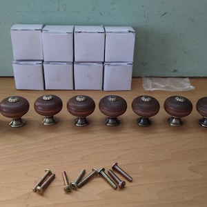 Könnte beinhalten: Acht braune Holzknauf für Schränke mit goldenen Akzenten und einer Reihe von Schrauben. Die Knäufe sind in einer Reihe auf einer hellbraunen Oberfläche angeordnet.