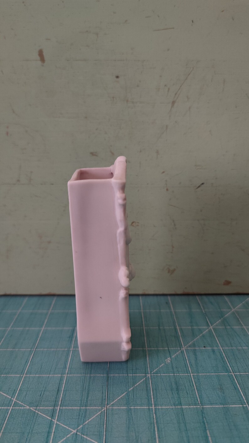 Puede incluir: Un jarr&oacute;n de cer&aacute;mica rosa p&aacute;lido con un dise&ntilde;o decorativo en el lateral. El jarr&oacute;n est&aacute; sobre una superficie de rejilla azul y blanca.