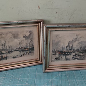May include: Two framed watercolor paintings depicting a harbor scene with ships and buildings. The paintings are in silver frames with a gold trim.