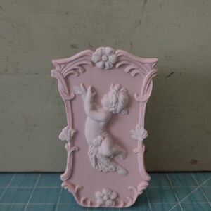 Puede incluir: Una placa de pared de cer&aacute;mica rosa con un marco ornamentado y una escultura de querub&iacute;n blanca en el centro.