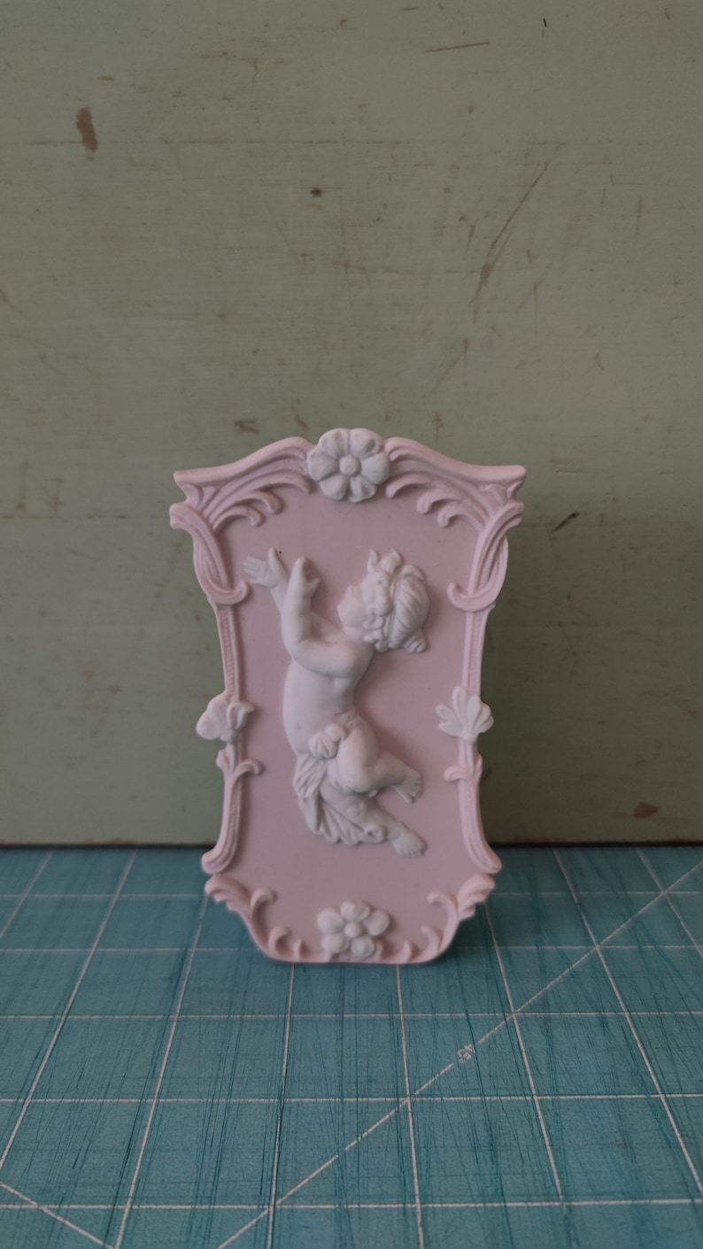 Puede incluir: Una placa de pared de cer&aacute;mica rosa con un marco ornamentado y una escultura de querub&iacute;n blanca en el centro.