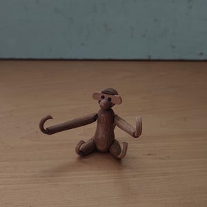 Könnte beinhalten: Kleine, handgefertigte Holz-Affenfigur mit ausgestreckten Armen und Beinen. Der Affe ist braun mit einem kleinen Hut und sitzt auf einer Holzoberfläche. Der Hintergrund ist eine hellblaue Wand.