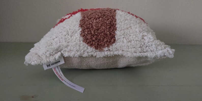 K&ouml;nnte beinhalten: Ein dekoratives Kissen mit einem strukturierten, getufteten Design. Das Kissen hat einen wei&szlig;en Hintergrund mit einem braunen Mittelstreifen und roten Akzenten. Es hat eine neutrale Stoffr&uuml;ckseite und ein Etikett.