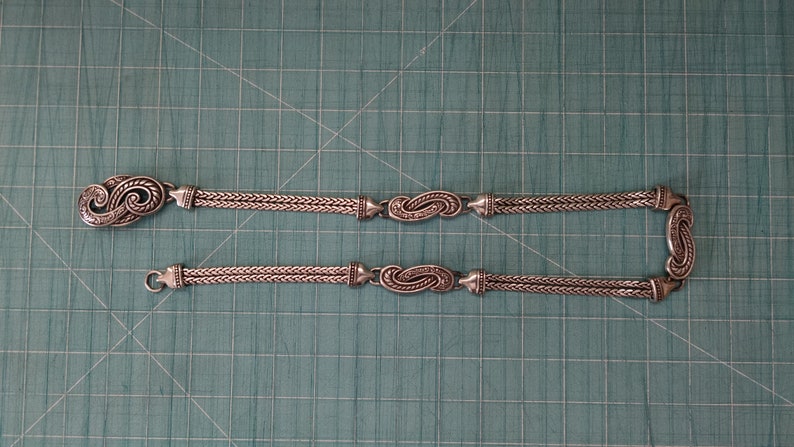 Peut inclure: Un collier en argent avec deux brins et des fermoirs en argent orn&eacute;s. Le collier est sur une surface verte.