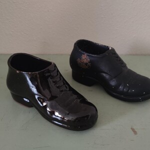Op de afbeelding: Twee zwarte keramische schoenen, een met een glanzende afwerking en de andere met een matte afwerking. Beide schoenen zijn met veters en hebben een klassiek ontwerp.