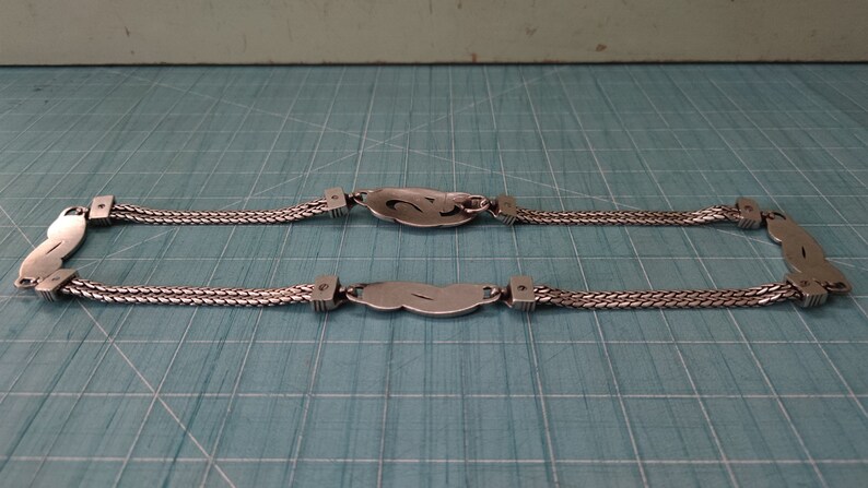 Peut inclure: Un collier en cha&icirc;ne argent avec deux pendentifs de forme ovale. Chaque pendentif pr&eacute;sente un motif stylis&eacute; et est reli&eacute; &agrave; la cha&icirc;ne par un petit anneau en argent.