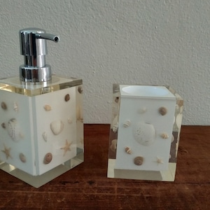 May include: A white soap dispenser and toothbrush holder set with a seashell design. The dispenser has a chrome pump and the holder has a clear top.