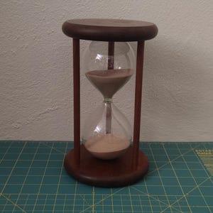 May include: An hourglass with a wooden frame and base. The clear glass bulb contains tan sand. The wooden frame is dark brown. The hourglass is a timekeeping device.