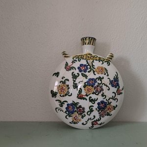 May include: A white ceramic vase with a round body and a floral design. The vase has a green, yellow, and blue floral pattern.